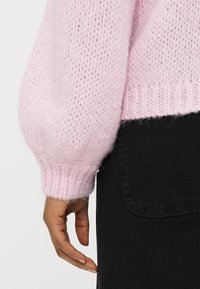 Χέρι χαλαρά στο πλάι, φορώντας ένα απαλό ροζ πουλόβερ με χοντρή πλέξη και μπαλόν μανίκι, και μαύρη φούστα από τζιν με εμφανή τσέπη.