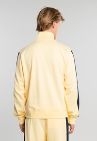 Hombre con chándal amarillo pálido con rayas negras en las mangas y los pantalones, de pie de espaldas contra un fondo gris.