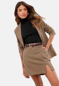 Braun gemusterter Blazer mit hochgerollten Ärmeln, schwarzem geripptem Rollkragenpullover und passendem gemustertem Rock mit seitlichem Schlitz und brauem Gürtel.