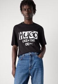 Black cotton t-shirt featuring a large white graphic with "HUGO" and "CREATIVE DEPT." Textured fabric, relaxed fit, round neckline.
