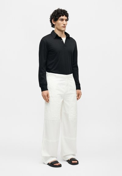 Άνδρας με σγουρά μαλλιά που φοράει μαύρο μακρυμάνικο πόλο, λευκό παντελόνι με φαρδιά μπατζάκια και μαύρα σανδάλια slide, στέκεται μπροστά σε απλό φόντο.
