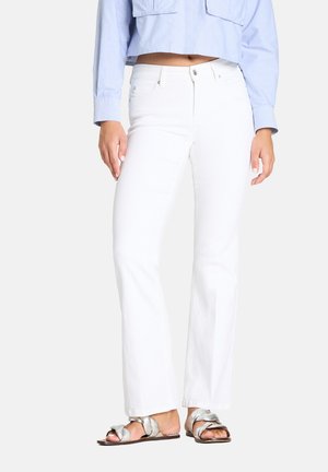 Woman wearing high-waist white jeans, light blue cropped shirt, and silver braided flat sandals, standing against a plain background.