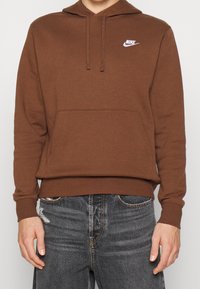 Hnědá mikina s klokaní kapsou, kapucí na šňůrku a bílým logem Nike na hrudi. Tkanina vypadá měkká a texturovaná.