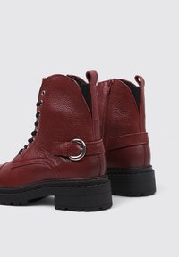 Botas de tobillo de cuero rojo con un acabado texturizado, cordones en la parte delantera, acento de anillo de metal y suela de goma negra y gruesa. Presenta un zipper lateral.