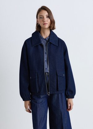 Junge Frau trägt dunkelblaue Jeansjacke und Hose über einem karierten Hemd, steht vor einem schlichten hellen Hintergrund.