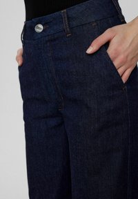 Dark blue denim pants with a straight cut, orange stitching, front pockets, and a silver button closure. Texture appears slightly brushed.