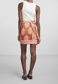 Falda envolvente naranja con patrón de paisley blanco, atada en la cintura, con un dobladillo texturizado. Usada con una camiseta sin mangas blanca y sandalias de tacón cuadrado negras.