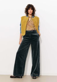 Gilet sans manches en polaire jaune sur un chemisier brun à motifs et un pantalon en velours côtelé large gris foncé. Des chaussures brunes complètent le look.