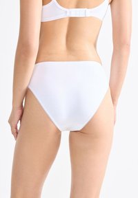 Donna che indossa biancheria intima bianca a vita alta e reggiseno bianco, vista di spalle su uno sfondo chiaro e uniforme.