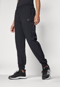 Črne atletske hlače z elastičnim pasom, stranskimi žepi in teksturiranimi črnimi stranskimi črtami. Ujemajo se s črnimi in belimi tekaškimi čevlji.