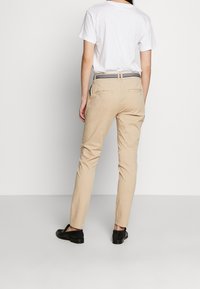 Chinos beiges coupe slim, avec une ceinture texturée et des poches arrière. Associés à un t-shirt blanc uni et des chaussures noires.