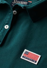 Polo shirt in diep groene katoen met een gestructureerde afwerking. Kenmerkt zich door een witte geborduurde patch met een vlagontwerp en de tekst "SUPERDRY".