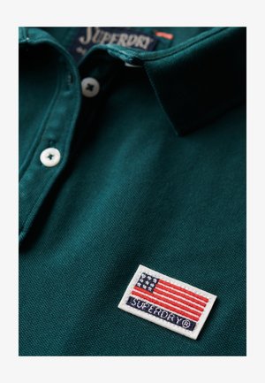 Polo shirt in diep groene katoen met een gestructureerde afwerking. Kenmerkt zich door een witte geborduurde patch met een vlagontwerp en de tekst "SUPERDRY".