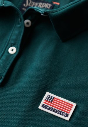 Polo en algodón verde oscuro con un acabado texturado. Presenta un parche bordado blanco con un diseño de bandera y el texto "SUPERDRY".