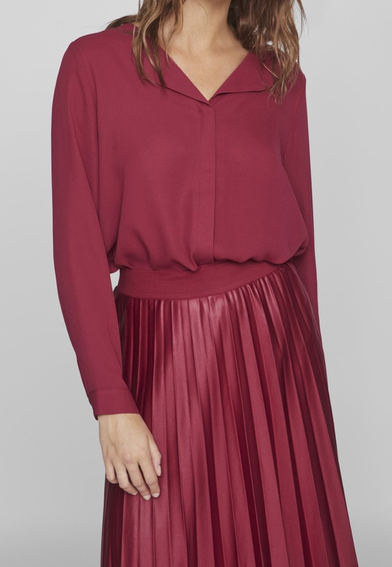 Femme portant un chemisier bordeaux à manches longues, rentré dans une jupe plissée bordeaux brillante, la main reposant le long du corps.