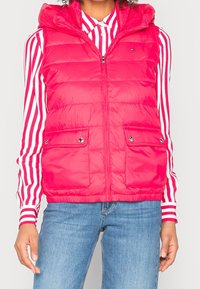 Gilet rose puffy avec capuche, doté d'une fermeture éclair à l'avant, de deux poches avec bouton, et d'une texture matelassée. Chemise rayée en dessous.