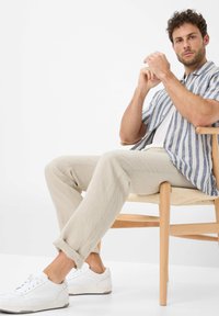 Blue and white striped short-sleeve shirt, beige linen pants, and white sneakers. Seated on a wooden chair with a woven seat.