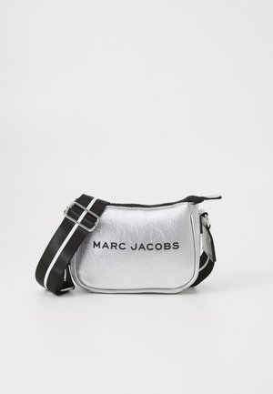 Hopeakylkylaukku, jossa on mustavalkoinen säädettävä olkahihna ja vetoketjusuljenta. Etupaneelissa on teksti "MARC JACOBS".