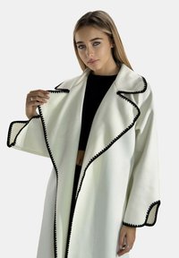 Hvid frakke med sort kant, der har en bred krave og et tekstureret stof. Designet inkluderer oversized ærmer og en åben front.
