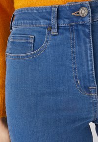 Jean en denim bleu avec une coupe slim, dotés d'une taille mi-haute, de coutures cuivres et d'une petite poche arrière. Tissu texturé avec un délavage minimal.