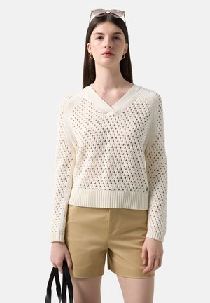 Jeune femme portant un pull en maille blanche à col en V, un short beige, des boucles d'oreilles créoles dorées, et tenant un sac cabas noir.