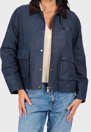 Frau trägt eine dunkelblaue Jacke mit messingfarbenen Druckknöpfen und Klappentaschen, kombiniert mit einem cremefarbenen Pullover und blauen Jeans.