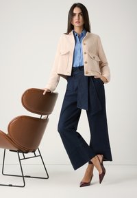Helle rosa Cord-Jacke mit zwei Fronttaschen, Knopfschluss; blaues Hemd; dunkelblaue weit geschnittene Hose mit Gürtel; burgunderfarbene spitze Absätze; brauner Stuhl.