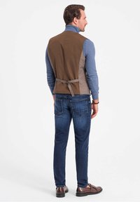 Weste aus braunem und grauem Mischgewebe mit einem Bindeband, getragen über einem blauen Rollkragenpullover, kombiniert mit dunklen Jeans und braunen Schuhen.