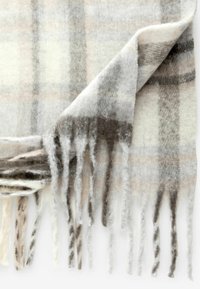 Écharpe en plaid en laine présentant des tons gris clair, beige et marron. Les bords sont ornés de franges, mettant en valeur une surface douce et texturée.