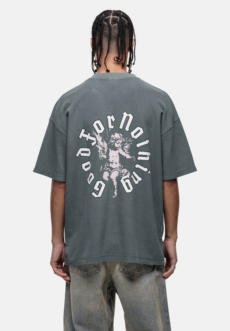 Zielony, oversize'owy t-shirt z fakturowanej tkaniny, na plecach z grafiką cheruba i napisem "Niezbyt Przydatny" w kontrastujących kolorach.