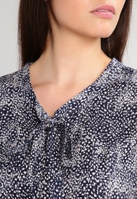 Granatowa bluzka w białe kropki, z dekoltem w kształcie litery V i zawiązanym węzełkiem przy kołnierzu. Materiał wydaje się lekki i teksturowany.