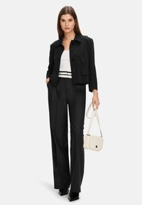 Black cropped jacket with buttons, white tank top, high-waisted black trousers, and a cream handbag. Model stands confidently.