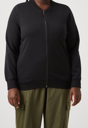 Person iført en sort lynlåstrøje med lange ærmer og olivengrønne cargobukser med klaplommer.