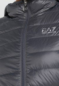 Chaqueta acolchada azul marino con cuello alto, cierre de cremallera y logo EA7 en relieve en el pecho, con textura suave y brillante.