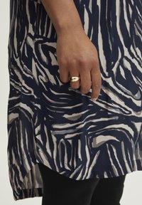 Marineblå tunika med beige abstrakte zebra-striber; letvægtsstof med en glat tekstur; båret med en gylden metallisk ring på hånden.