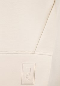 Tejido de sudadera beige con una textura suave, que presenta una costura vertical y un parche de logo rectangular. Diseño minimalista con líneas limpias.