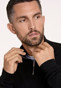 Felpa nera con zip realizzata in un materiale morbido, con una zip argentata e polsini a costine. Il modello sistema il colletto, mettendo in evidenza la texture e la vestibilità.