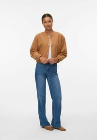Femme se tenant debout dans une veste beige, un haut blanc, un jean bleu et des chaussures à talons pointus marron sur un fond blanc uni.