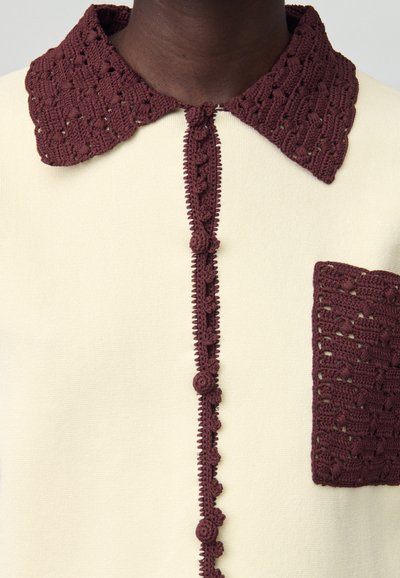 Suéter crema con un cuello de ganchillo burdeos texturizado y un bolsillo, que presenta un panel frontal decorativo con botones y un intrincado detalle.