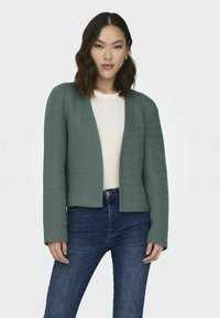 Groene gestructureerde blazer met een cropped ontwerp, open voorkant en twee voorkeurszakken, gedragen over een lichtgekleurde top en donkere jeans.