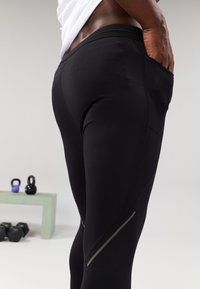 Juodos sportinės legginsus su prigludusia forma, šonuose turinčiais kišenes ir atspindinčius akcentus palei siūles. Lygus paviršius ir elastinga medžiaga.