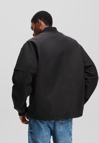 Jachetă bomber neagră cu guler înalt, finisaj mat texturat și siluetă spațioasă. Include mâneci raglan și manșete elastice.