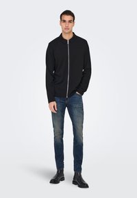 Melna jaka ar rāvējslēdzēju un apkakli, kopā ar zilām džinsa biksēm. Modelis stāv melnos zābakos, demonstrējot minimālistisku dizainu un piegriezumu.