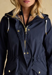 Frau trägt eine dunkelblaue Kapuzenjacke mit goldfarbenem Reißverschluss und Druckknöpfen, geschichtet über einem beige und dunkelblauen Pullover.
