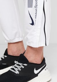 Osoba v bílých teplácích se značkou Nike a černých teniskách Nike Flyknit s potištěnými tkaničkami a květinovým vzorem na patě.