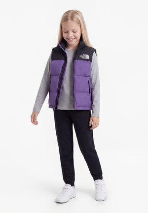 Dieťa s dlhými blond vlasmi, ktoré má na sebe fialovú a čiernu vestu North Face, sivé tričko s dlhým rukávom, čierne nohavice a biele tenisky, sa usmieva a pozerá dolu.