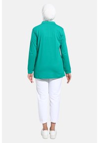 Maglia a maniche lunghe color turchese in tessuto morbido, con collo a camicia, abbinata a pantaloni bianchi alla caviglia e sneakers bianche.