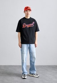 Camiseta negra de manga corta con el logo "Dsquared²" en rojo y azul, combinada con jeans de mezclilla azul claro y zapatillas en blanco y negro. Lleva una gorra roja.