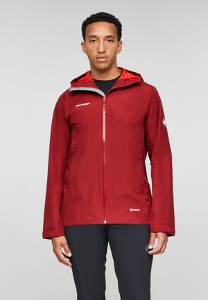 Uomo con giacca rossa Mammut Gore-Tex con cappuccio e pantaloni neri, in piedi davanti a uno sfondo chiaro uniforme.