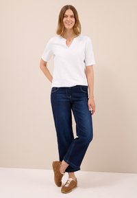Kvinde står barfodet på ét ben, iført hvid kortærmet skjorte, mørkeblå brede jeans og brune sneakers, smilende mod en beige baggrund.